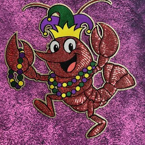 May include: A red sequined crawfish cartoon character wearing a jester hat and a beaded necklace. The crawfish has a smiling face and is holding a strand of beads in its claw. The background is a textured purple.