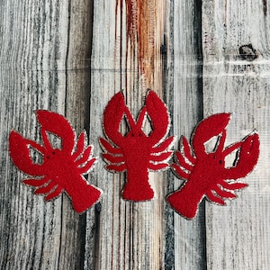 May include: Three red felt lobster appliqués with silver glitter trim are arranged on a weathered wood-grain background. The lobsters are detailed with claws, eyes, and segmented bodies, perfect for crafting or nautical-themed projects.