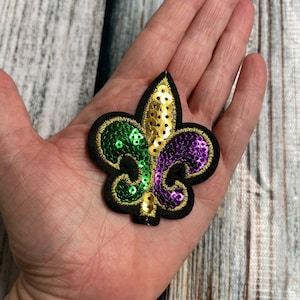 May include: A sequined fleur-de-lis patch in green, gold, and purple, with a black border. The patch is held in a hand, showcasing the intricate details of the sequins and the design's shape. The patch is a symbol of New Orleans.