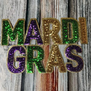May include: Mardi Gras themed patch with the words "Mardi Gras" in gold, green, and purple sequins.