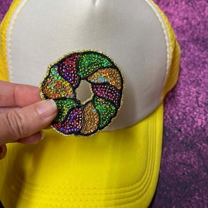 May include: A yellow and white trucker hat with a colorful sequined patch in the shape of a circle. The patch has a gold border and features green, purple, red, and gold sequins. The hat is made of fabric and mesh.