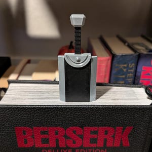 Puede incluir: Réplica en miniatura de una espada, con una combinación de colores gris y negro. La espada está colocada encima de un libro con la palabra "BERSERK" en letras rojas. El fondo incluye otros libros.