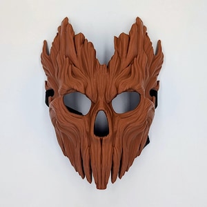 May include: A brown mask with a textured surface resembling tree bark. The mask features deep vertical grooves, a pointed top edge, two large eye openings, and a central nose cavity. A black elastic strap is visible on the sides for securing it.
