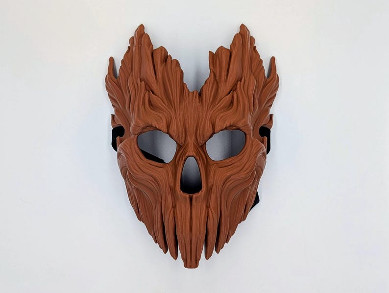 May include: A brown mask with a textured surface resembling tree bark. The mask features deep vertical grooves, a pointed top edge, two large eye openings, and a central nose cavity. A black elastic strap is visible on the sides for securing it.