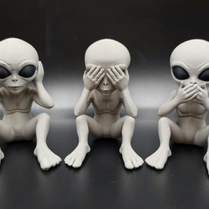 May include: Three gray alien figurines in the "See No Evil, Hear No Evil, Speak No Evil" pose. Each figurine has large black oval eyes and is seated with hands covering the eyes, ears, and mouth. The figurines are on a dark surface.