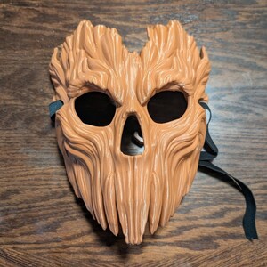 May include: A light brown mask with a textured surface resembling tree bark. The mask features deep, vertical grooves, two large eye openings, and a triangular nose opening. A black elastic strap is attached to the sides for securing the mask.