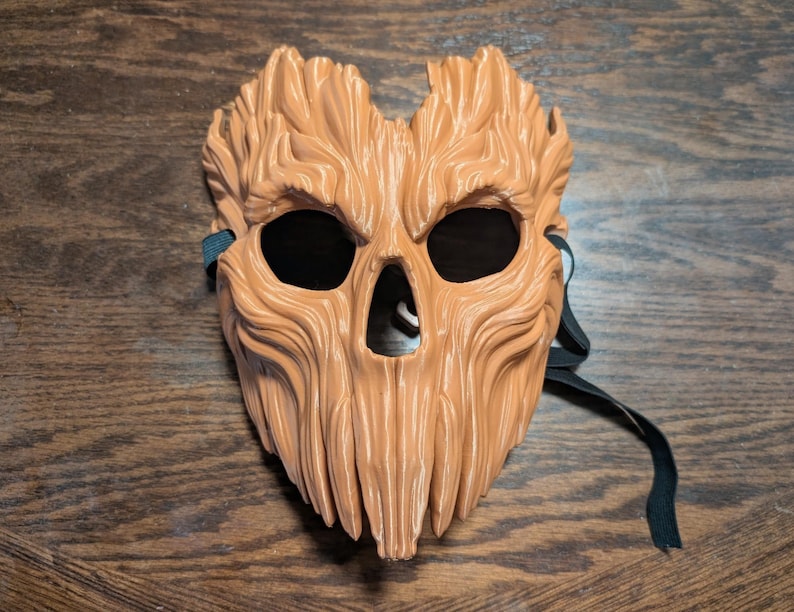 May include: A light brown mask with a textured surface resembling tree bark. The mask features deep, vertical grooves, two large eye openings, and a triangular nose opening. A black elastic strap is attached to the sides for securing the mask.