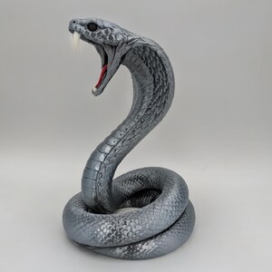 Puede incluir: Una figura de cobra gris en posición de ataque, enroscada y erguida, con la boca abierta mostrando un interior rojo y colmillos blancos. La serpiente presenta escamas detalladas en todo el cuerpo, un ojo oscuro y un acabado suave y texturizado.