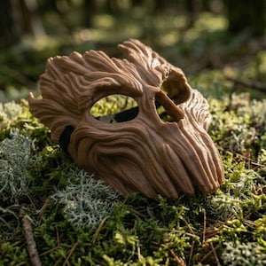 May include: A brown mask with a textured surface resembling tree bark, featuring deep grooves, a prominent brow, and two large eye openings. The mask has a rigid, organic shape with a jagged top edge and a black elastic strap visible on the side.