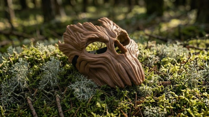 May include: A brown mask with a textured surface resembling tree bark, featuring deep grooves, a prominent brow, and two large eye openings. The mask has a rigid, organic shape with a jagged top edge and a black elastic strap visible on the side.