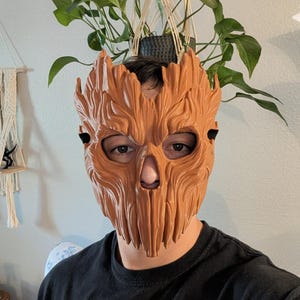 May include: A light brown, textured mask with a wood-grain appearance and vertical grooves, featuring cutouts for the eyes and nose. The mask covers the upper face and is secured with black straps, displaying a detailed, organic, bark-like surface.