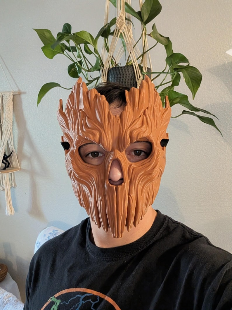 May include: A light brown, textured mask with a wood-grain appearance and vertical grooves, featuring cutouts for the eyes and nose. The mask covers the upper face and is secured with black straps, displaying a detailed, organic, bark-like surface.