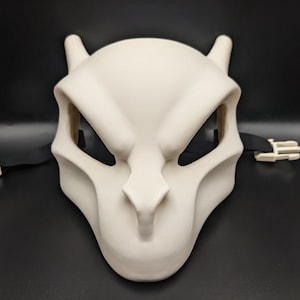 May include: A smooth, off-white, molded mask featuring a pointed snout, angular eye openings, and two small horn-like protrusions at the top. The mask includes black elastic straps on both sides, each finished with a white plastic side-release buckle.