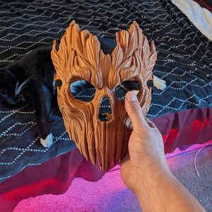May include: A brown, textured mask with a wood-like appearance, featuring deep grooves, carved details, eye openings, and a nose cavity. The mask has a jagged top edge and includes a black elastic strap attached to the sides for securing it in place.