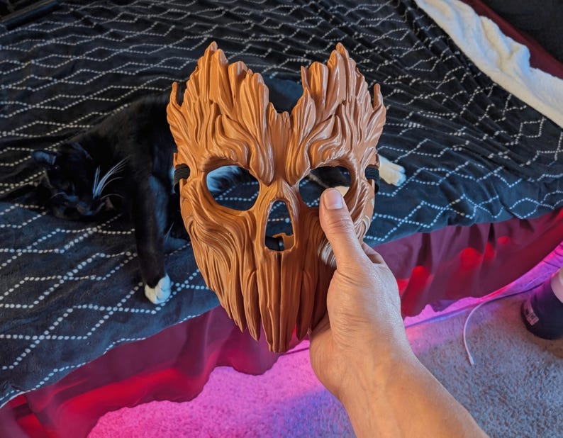 May include: A brown, textured mask with a wood-like appearance, featuring deep grooves, carved details, eye openings, and a nose cavity. The mask has a jagged top edge and includes a black elastic strap attached to the sides for securing it in place.