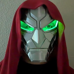 May include: A metallic silver face mask with angular, geometric plating and bright green LED lights glowing from the eye sockets. The mask is framed by a deep red fabric hood that drapes over the head and shoulders, creating a stark contrast.