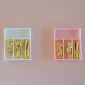 May include: Two small, rectangular containers with compartments, each holding gold-colored hair pins. One container is white, the other is pink. The background is a solid, light pink color.