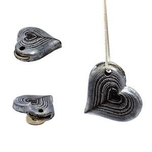 Puede incluir: Un colgante de corazón plateado con un patrón de rayas blancas y negras cuelga de una cadena plateada. El colgante tiene un pequeño agujero en la parte superior para sujetar la cadena.