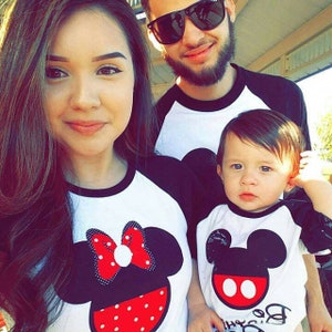 May include: A family of three wearing matching Mickey Mouse themed shirts. The shirts are white with black sleeves and feature a large Mickey Mouse head with red polka dots. The woman is wearing a red bow on her Mickey Mouse head. The man is wearing a black baseball cap. The child is wearing a black and white Mickey Mouse head with red polka dots and the text "Be Our Guest" on the bottom.