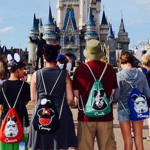 May include: A group of people standing in front of Cinderella's Castle at Walt Disney World. They are all wearing drawstring bags with different Disney characters on them. The bags say "Golden", "Tammie", "Chris", and "Kinsey" on them.