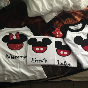 May include: Four white t-shirts with black and red Mickey Mouse designs. The shirts have the names "Mommy", "Stevie", "Julian" and "Pixie" printed on them.