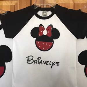 May include: White and black raglan t-shirt with a red and white polka dot Minnie Mouse head graphic and the name "BRIANELPS" printed in black below.