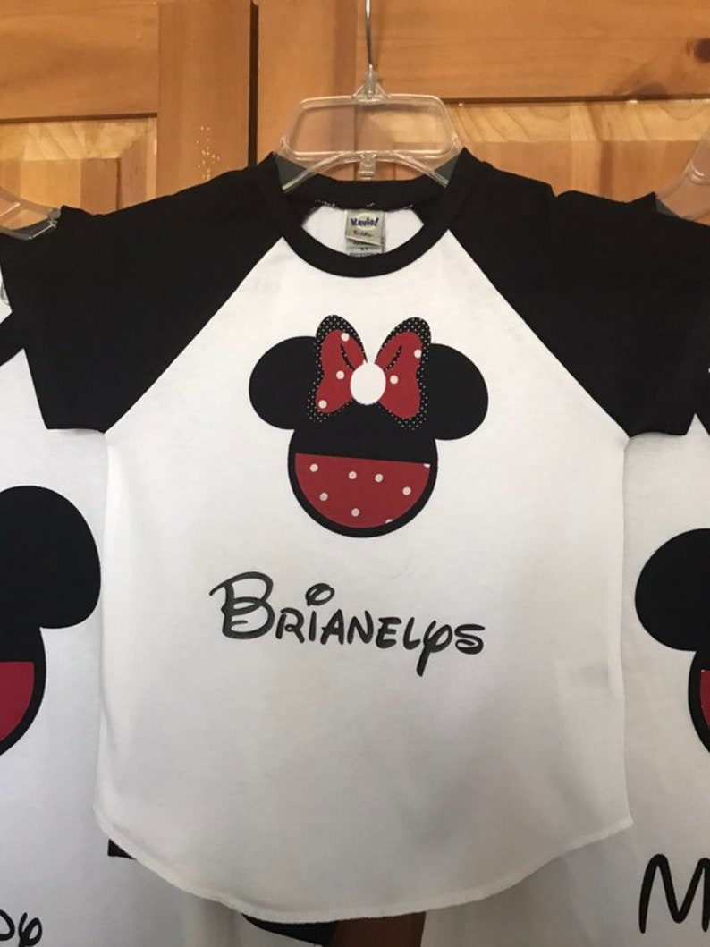 May include: White and black raglan t-shirt with a red and white polka dot Minnie Mouse head graphic and the name "BRIANELPS" printed in black below.