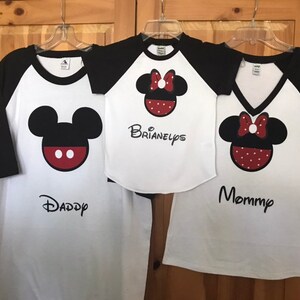 May include: Three white t-shirts with black sleeves and a Mickey Mouse or Minnie Mouse design. The shirts say "Daddy", "Mommy", and "Brianelyps".