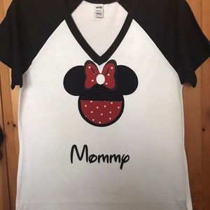 May include: White t-shirt with black sleeves and a black v-neck. The shirt features a red and white polka dot Minnie Mouse head with a red bow and the word "Mommy" in black below it.
