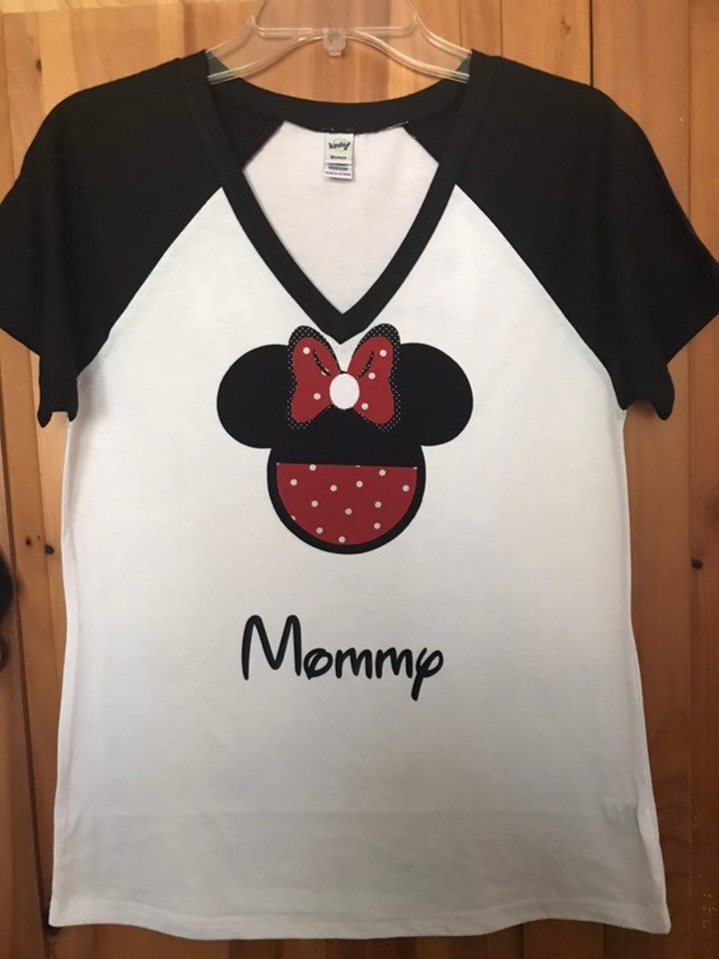 May include: White t-shirt with black sleeves and a black v-neck. The shirt features a red and white polka dot Minnie Mouse head with a red bow and the word "Mommy" in black below it.