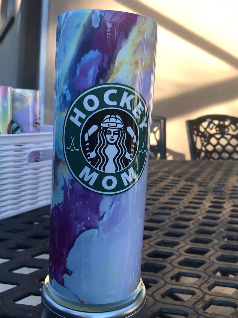 May include: A stainless steel tumbler with a marbleized purple, blue, and yellow design. The tumbler has a green and white logo that says "Hockey Mom" with a hockey stick and a woman's face wearing a hockey helmet.
