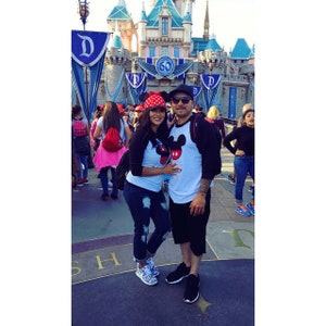 May include: A couple wearing matching Mickey Mouse shirts with red hats stand in front of Sleeping Beauty's castle at Disneyland. The man is wearing a white shirt with a black baseball jersey over it. The woman is wearing a black and white striped shirt. The castle is decorated with flags and banners.