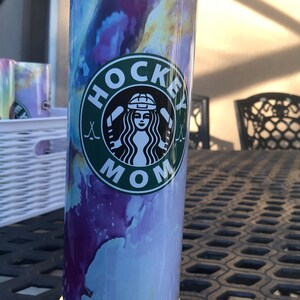 May include: A stainless steel tumbler with a purple, blue, and yellow watercolor design. The tumbler has a green circle with the words "HOCKEY MOM" and a hockey stick graphic.