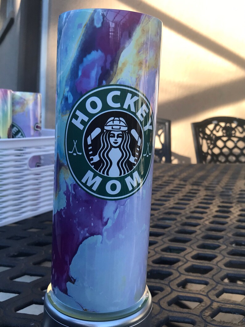 May include: A stainless steel tumbler with a purple, blue, and yellow watercolor design. The tumbler has a green circle with the words "HOCKEY MOM" and a hockey stick graphic.