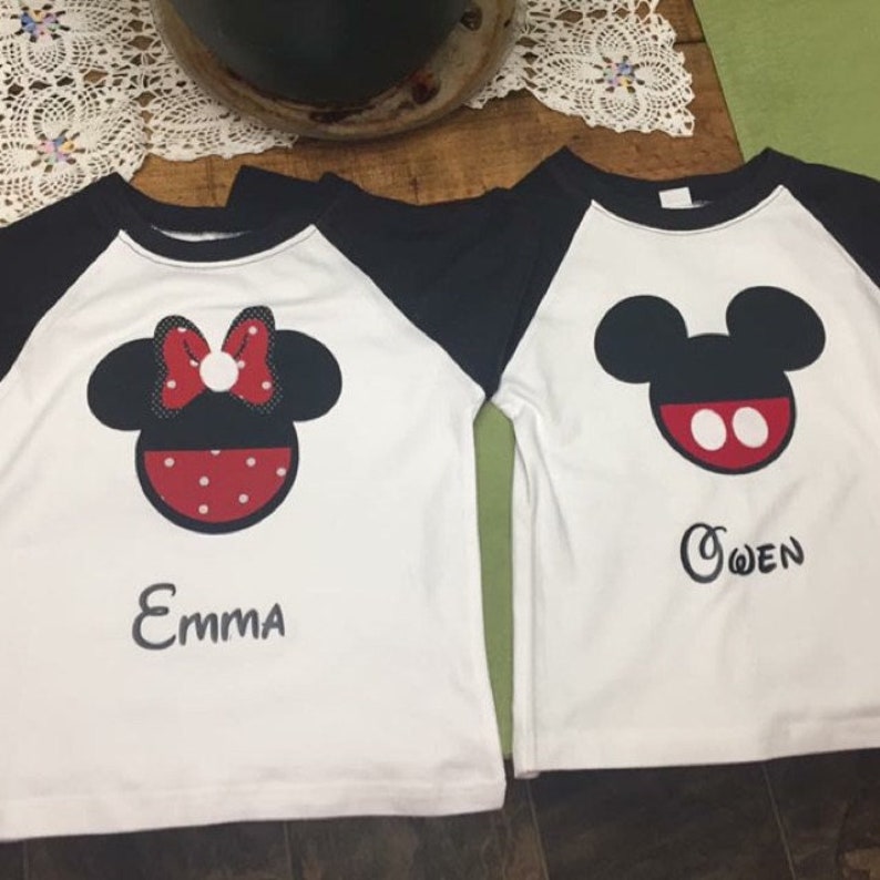 May include: Two white t-shirts with black sleeves. One shirt features a red and white Minnie Mouse design with the name "Emma" printed below. The other shirt features a black Mickey Mouse design with the name "Owen" printed below.