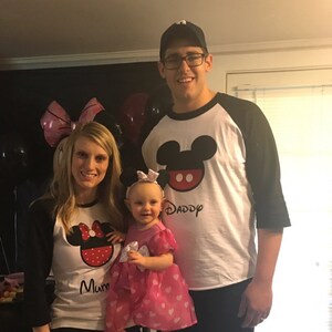 May include: A family of three wearing matching Mickey Mouse themed shirts. The father is wearing a black and white baseball shirt with Mickey Mouse's head and the word "Daddy" on it. The mother is wearing a black and white baseball shirt with Minnie Mouse's head and the word "Mum" on it. The child is wearing a pink dress with Minnie Mouse's head on it.