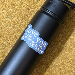 May include: A black reusable water bottle with a white sticker that says "Love you to the moon & to saturn" with a crescent moon and a planet with rings.