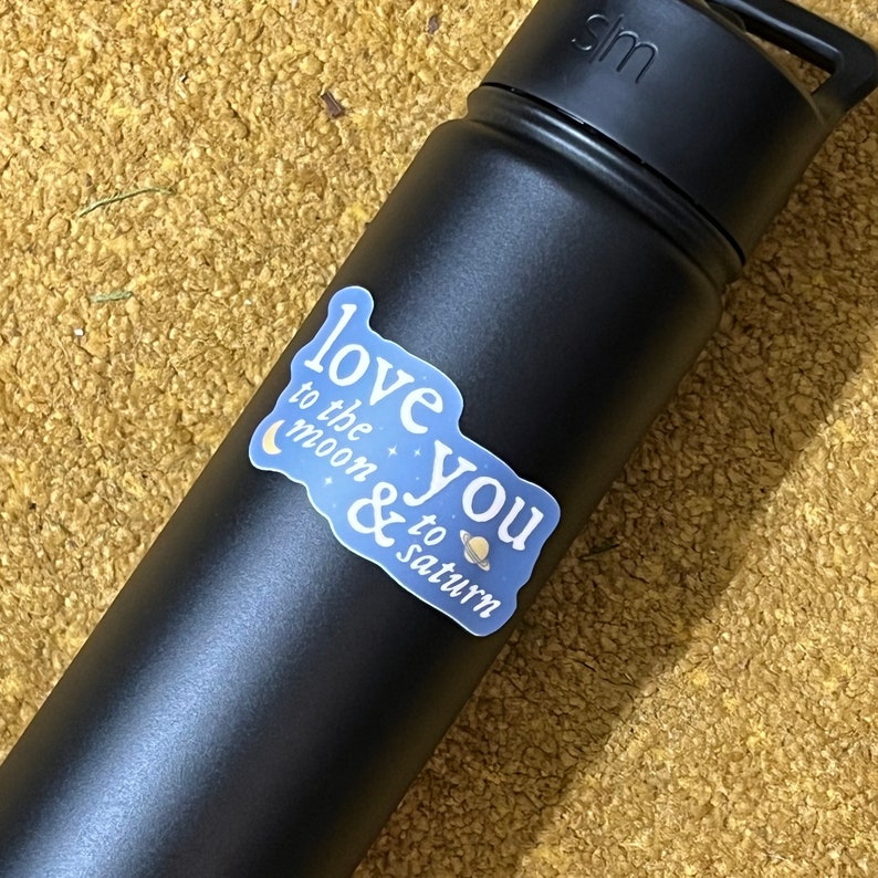 May include: A black reusable water bottle with a white sticker that says "Love you to the moon & to saturn" with a crescent moon and a planet with rings.