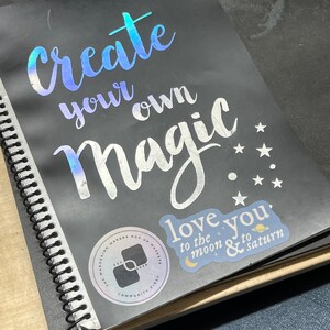 May include: A black spiral bound notebook with a silver holographic sticker that says "create your own magic." There is a smaller sticker that says "love you to the moon & to saturn" with a crescent moon and a planet graphic. The notebook also has a round sticker with the text "The Wandering Makers Pop Up Market Est. 2020 Community First."