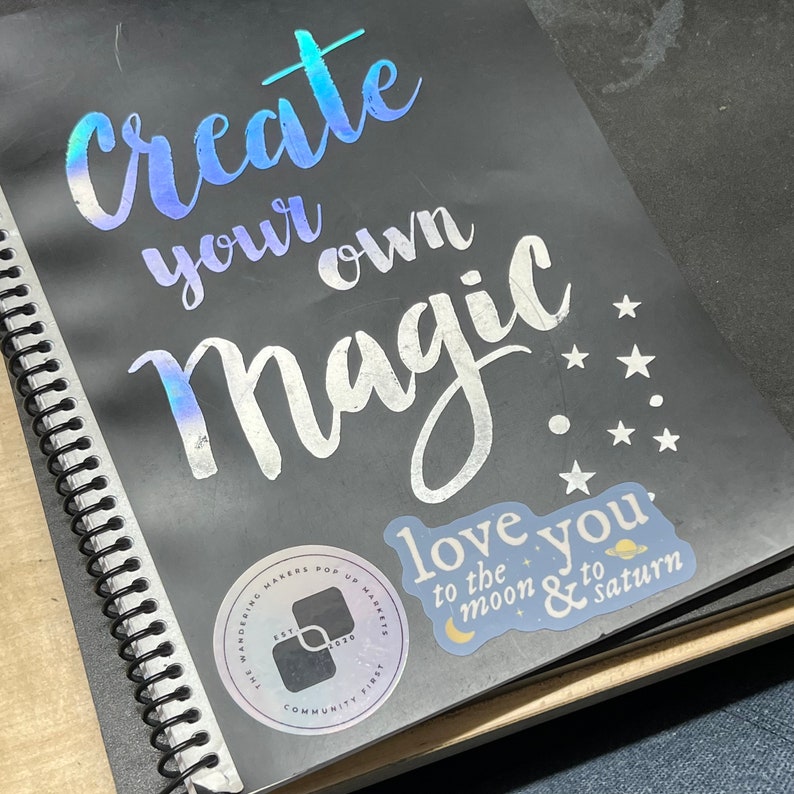 May include: A black spiral bound notebook with a silver holographic sticker that says "create your own magic." There is a smaller sticker that says "love you to the moon & to saturn" with a crescent moon and a planet graphic. The notebook also has a round sticker with the text "The Wandering Makers Pop Up Market Est. 2020 Community First."