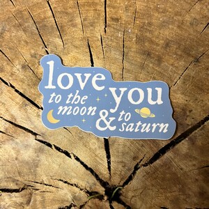 May include: A blue sticker with white text that reads "love you to the moon & to saturn". The sticker features a crescent moon and a planet with rings.