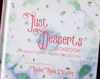 May include: A white cookbook with a watercolor design on the cover. The title is "Just Desserts Cookbook" and the subtitle is "A Stanley Family Recipe Collection". The author is Rachel Alana Stanley.