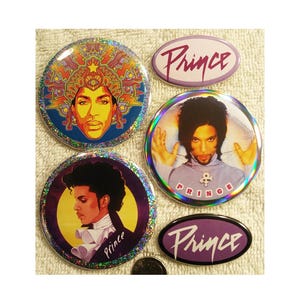 May include: A collection of four circular and two oval Prince buttons. The circular buttons feature images of Prince with various designs and colors. The oval buttons have the name "Prince" in a stylized font. The buttons have a holographic, glittery effect.