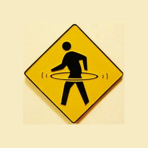 May include: A yellow diamond-shaped sign with a black border. The sign features a black silhouette of a person hula hooping. The hula hoop is a light gray color. The background is a light beige.