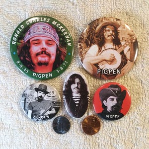 May include: A collection of vintage pinback buttons featuring images of Ronald "Pigpen" McKernan. The buttons vary in size and design, with some including text such as "Pigpen" and dates. The buttons are in various colors, including green, red, and sepia.