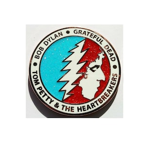 May include: A round pin with a copper-colored border. The pin features a blue glitter background, a white lightning bolt, and a red background with a white profile. The pin has text that reads "BOB DYLAN", "GRATEFUL DEAD", and "TOM PETTY & THE HEARTBREAKERS".