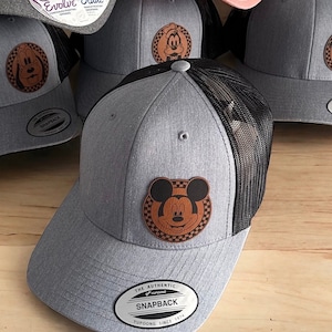 May include: Six gray and pink baseball caps with black mesh back panels. Each cap features a brown leather patch with a cartoon character on it. The patches feature Mickey Mouse, Minnie Mouse, Pluto, and Goofy. The caps have the text "Evolve Eludes" and "The Authentic Snapback" on them.