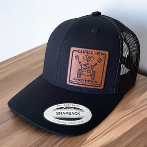 May include: Black and brown trucker hat with a leather patch featuring a cartoon robot and the text "WALL-E" and "Earthside Artifacts & Antiques". The hat has a black mesh back and a black snap closure with the text "The Authentic Yupoong Snapback Yupoong Since 1974".