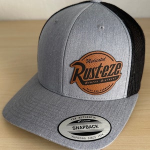 May include: Gray and black baseball cap with a brown leather patch that says "Medicated Rust-eze Bumper Ointment Rear End Formula". The cap has a silver sticker that says "The Authentic YUPOONG Snapback YUPOONG Since 1974".