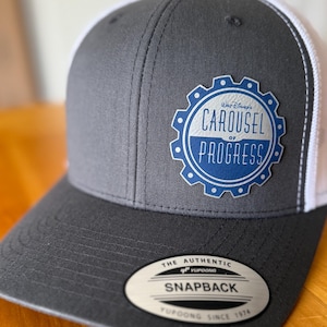 May include: A gray and white snapback hat with a curved brim. The hat features a circular patch with the text "Walt Disney's Carousel of Progress" in blue and white. The hat has a snap closure and a Yupoong tag.